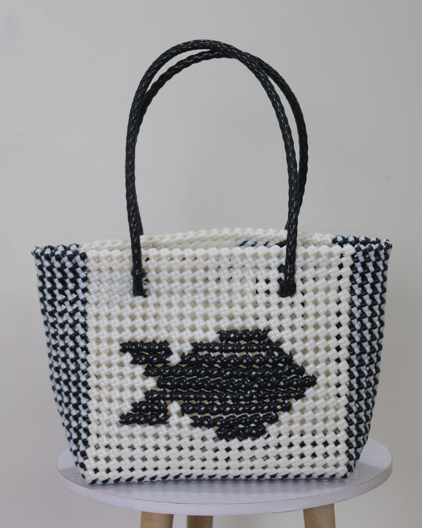 Black and white knitted handbag with a fish knitted design in the middle of it, place on a white stool against a light gray background