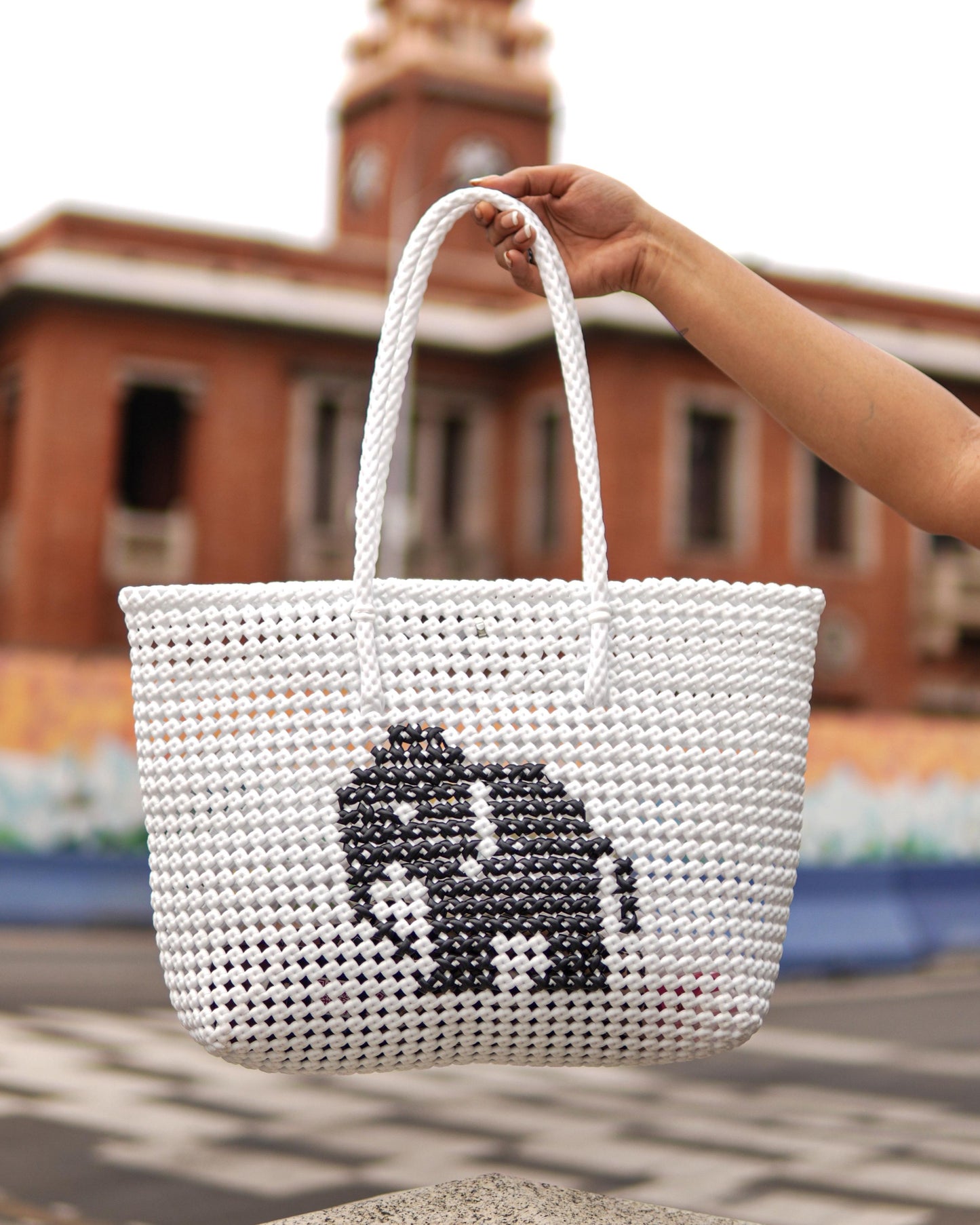 White knitted handbag with an elephant knitted design in the middle, held by a hand in front of a blurred building.
