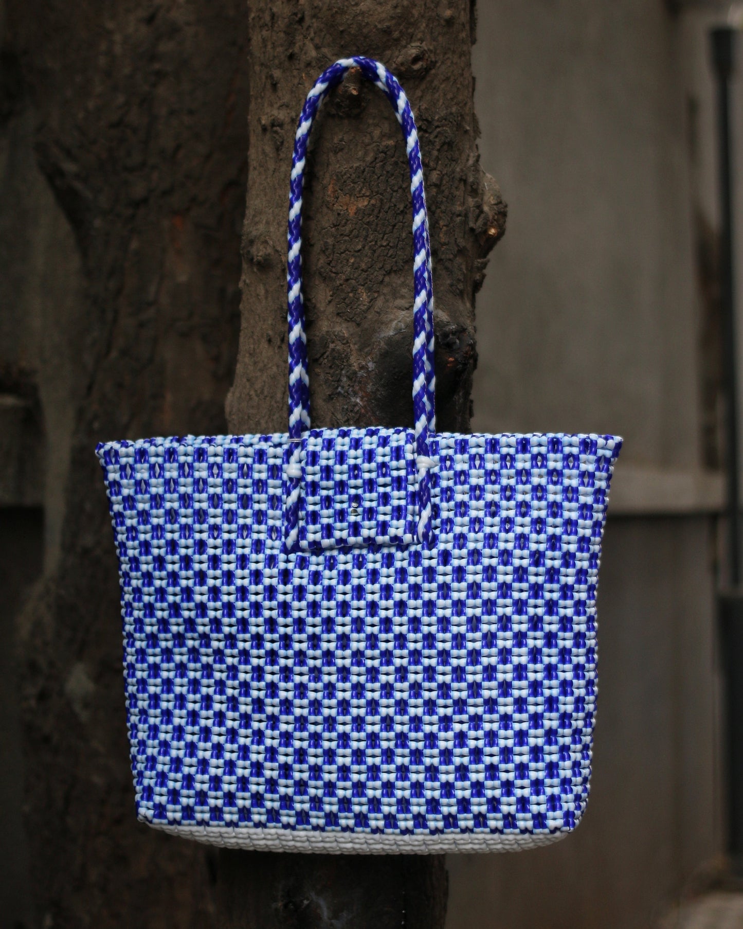 Blue and white knitted handbag with braided handle hanging from a tree bark.