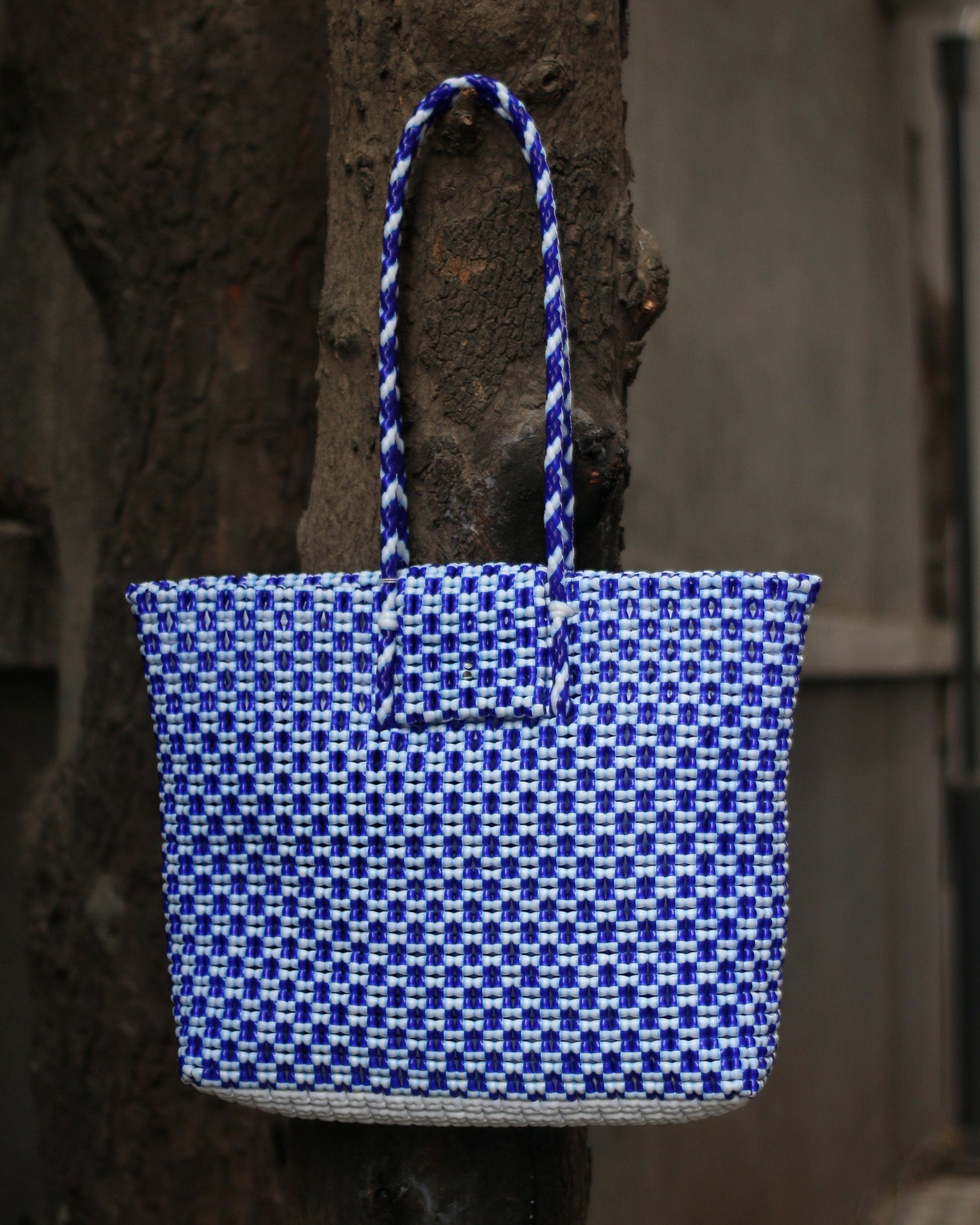 Blue and white knitted handbag with braided handle hanging from a tree bark.
