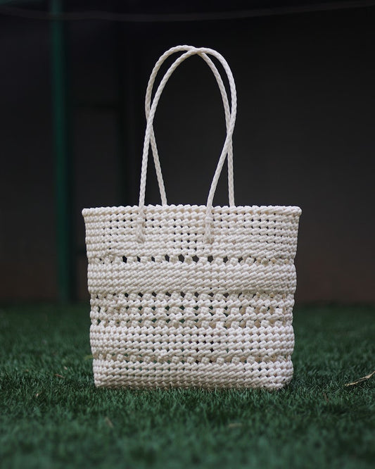 White knitted handbag with braided handle, placed on a grass floor with blurred background.