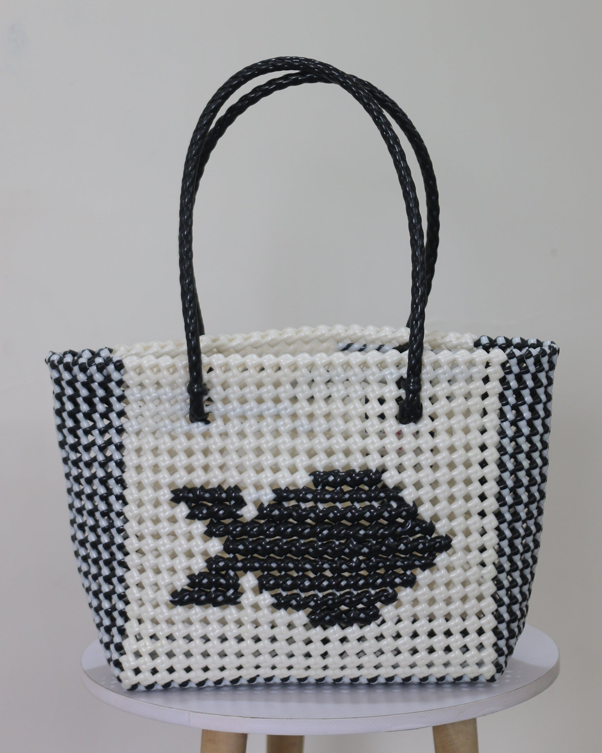 Black and white knitted handbag with a fish knitted design in the middle of it, place on a white stool against a light gray background