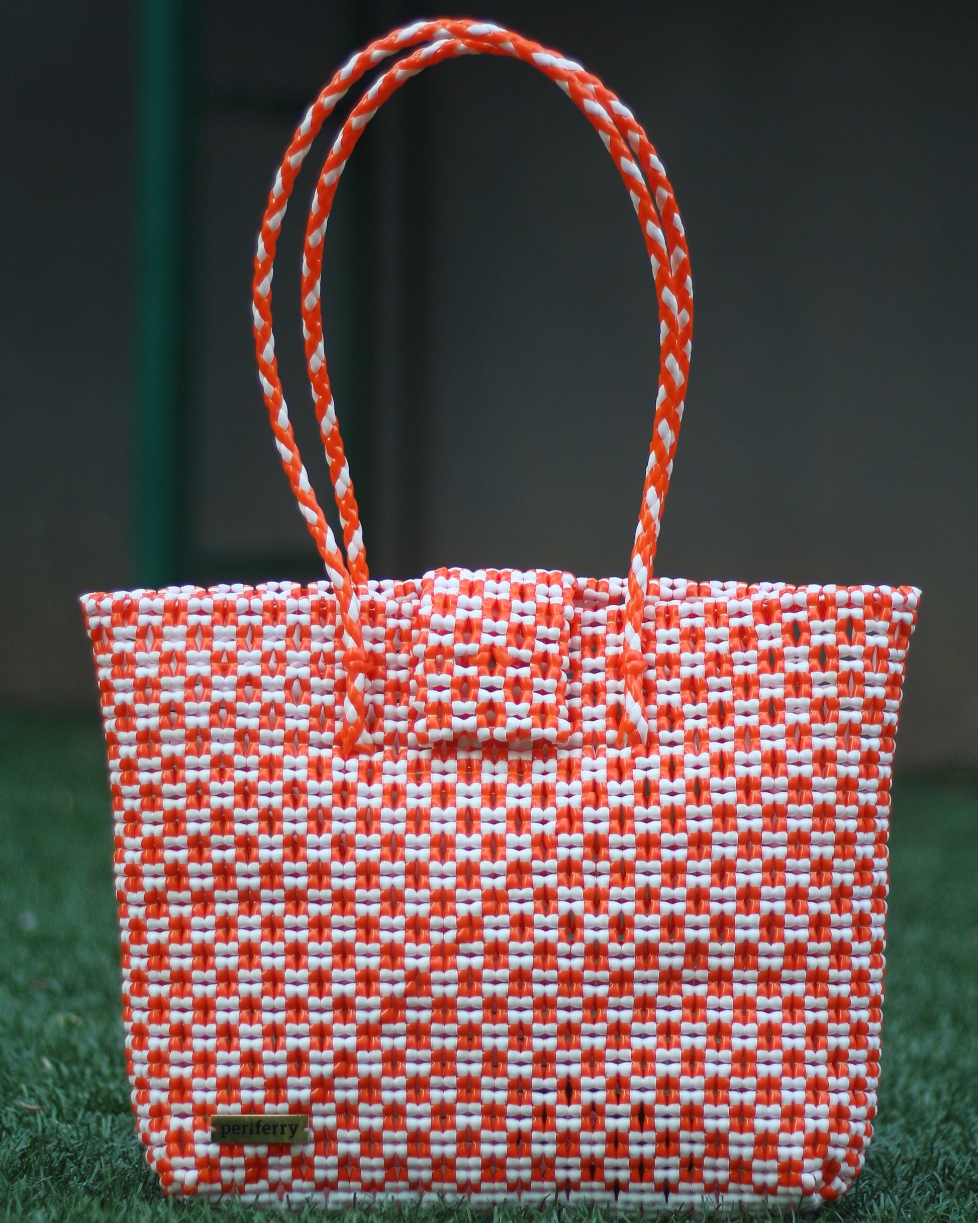 Red and white knitted handbag with braided handles on a grassy surface.