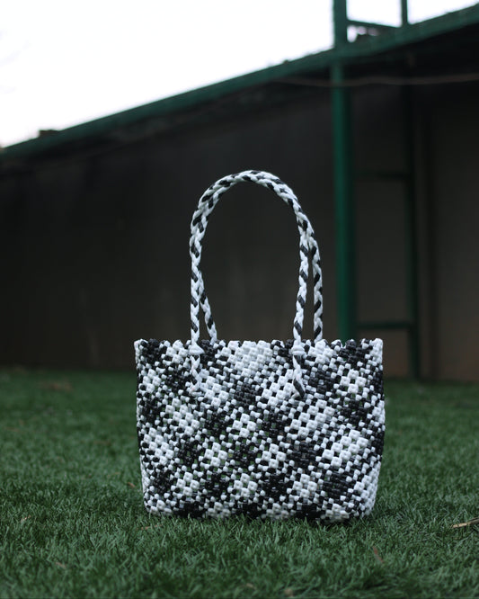 Black and White knitted in a checkered pattern handbag with braided handles, placed on a grass surface with a blurred background.