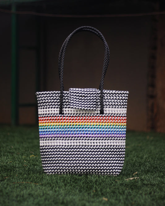 Black and white knitted handbag with rainbow themed knitting in the middle, with black braided handle, placed on grass surface with blurred background.
