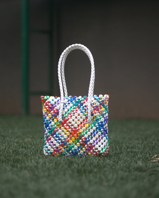 A small, rainbow colored knitted handbag with long braided handle, placed on a grass floor with blurred background.