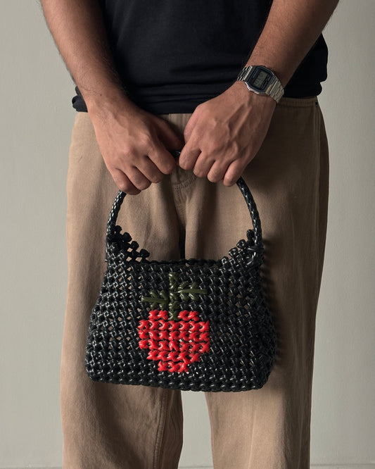 Person holding a black knitted handbag with a red apple design in the middle, on a neutral background