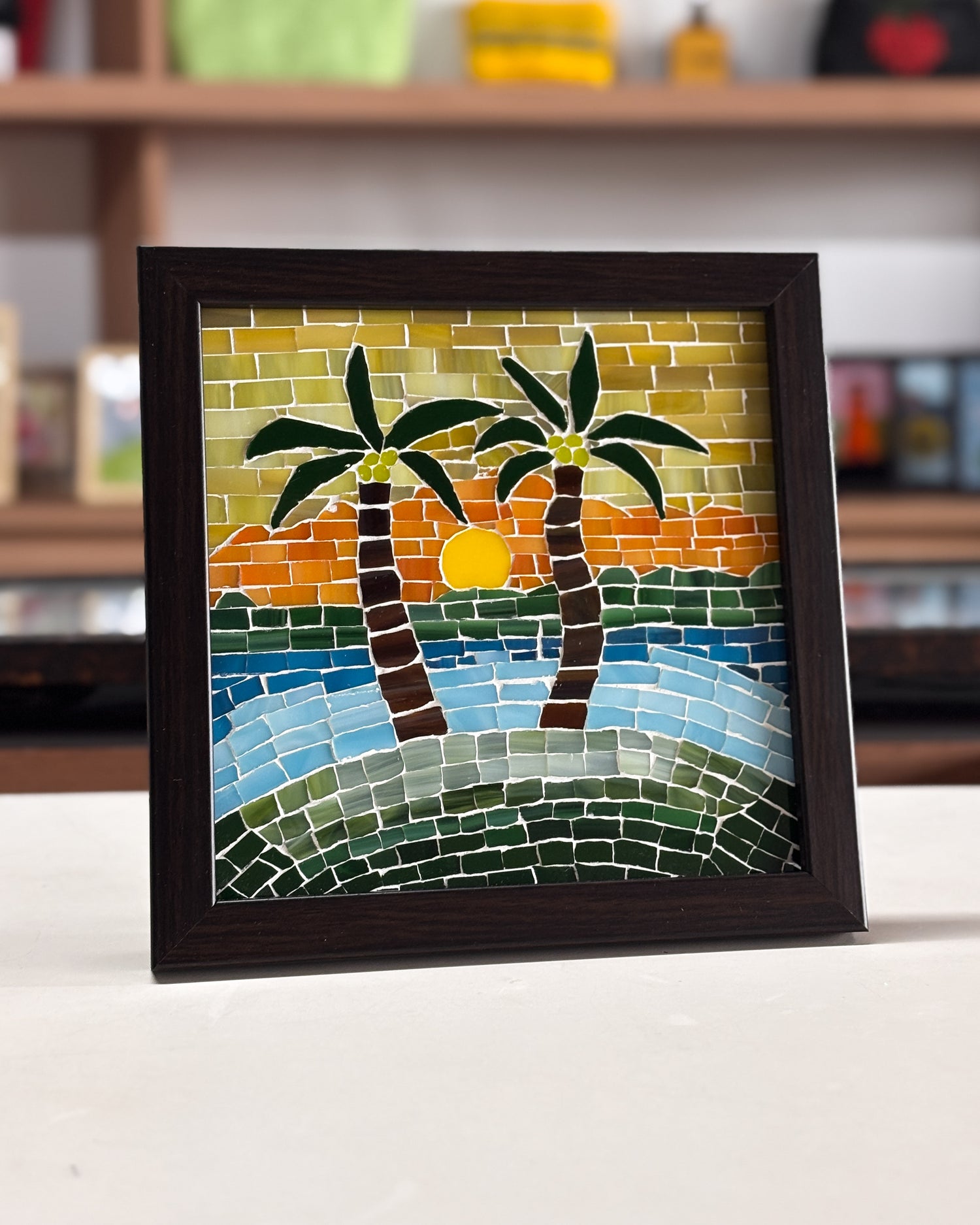Framed mosaic artwork of palm trees and a sunset on a table.