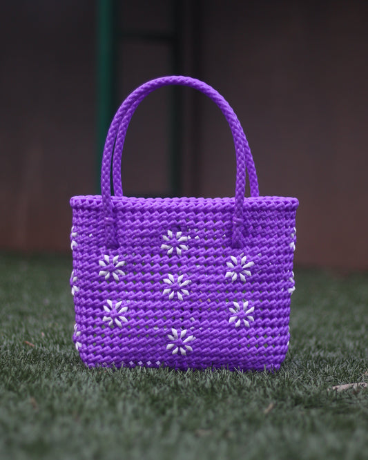 Purple knitted handbag with white knitted floral design all over it, with a braided handle placed on a grass floor with blurred background.